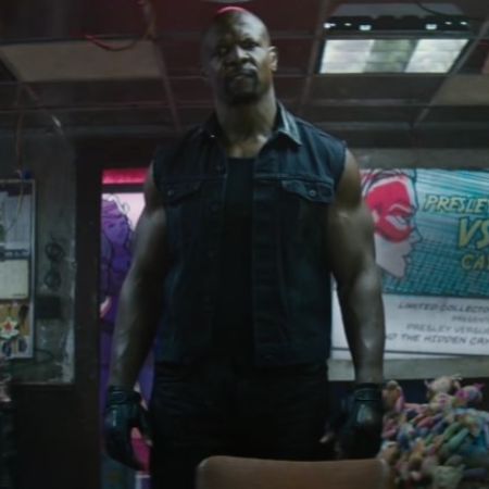 Terry Crews is standing aggressively wearing a sleeveless jacket with black gloves on. 